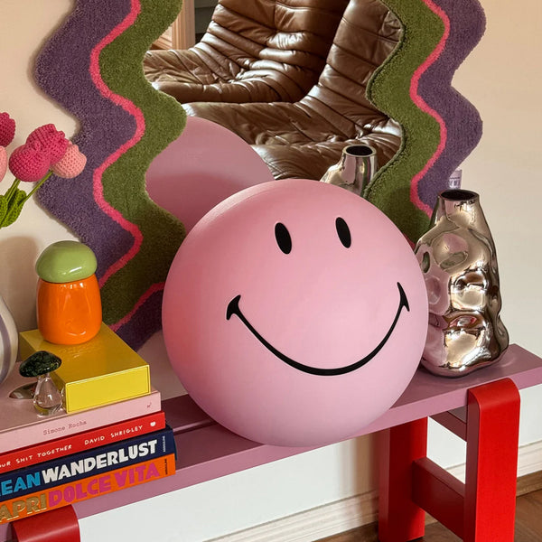 Pink smiley face ball on a shelf with decorative items and books.